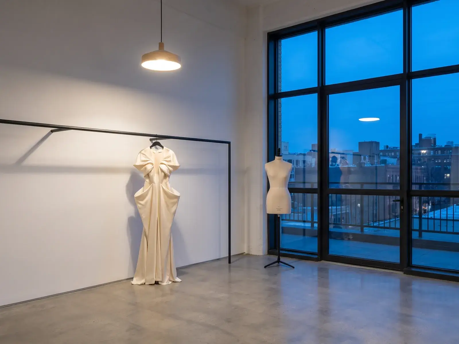 A finished gown on a rail in the atelier at dusk, city windows behind it