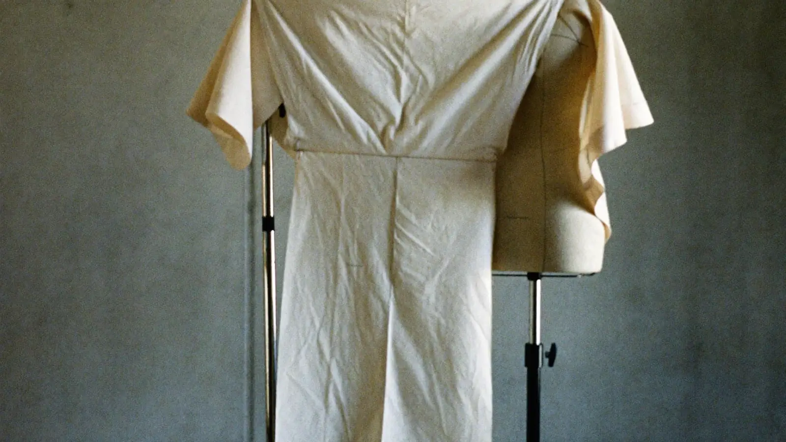 A cotton toile pinned to a dress stand against a bare concrete wall, hard side light raking across the seams, tailor's chalk marks and pins visible, muted grey and bone tones, one corner of the cloth lifted by movement.