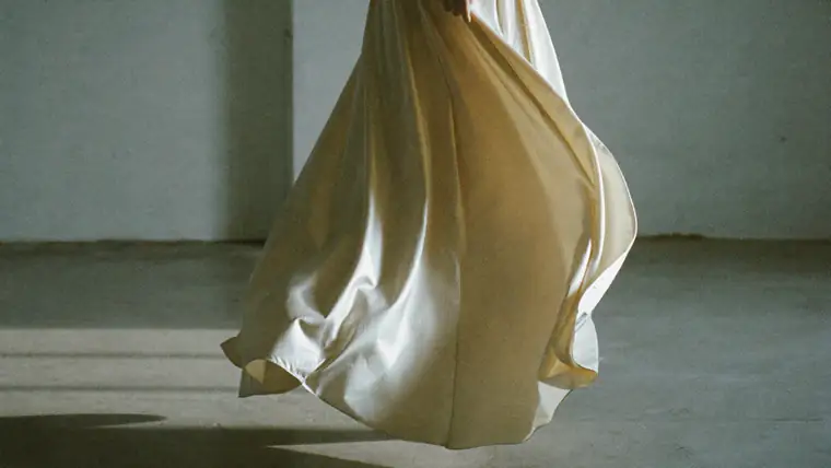 A long ivory silk skirt caught mid-swing in a bare atelier, hard side light from a tall window, raw concrete floor, a paper calendar page pinned to the wall behind, muted bone and grey.