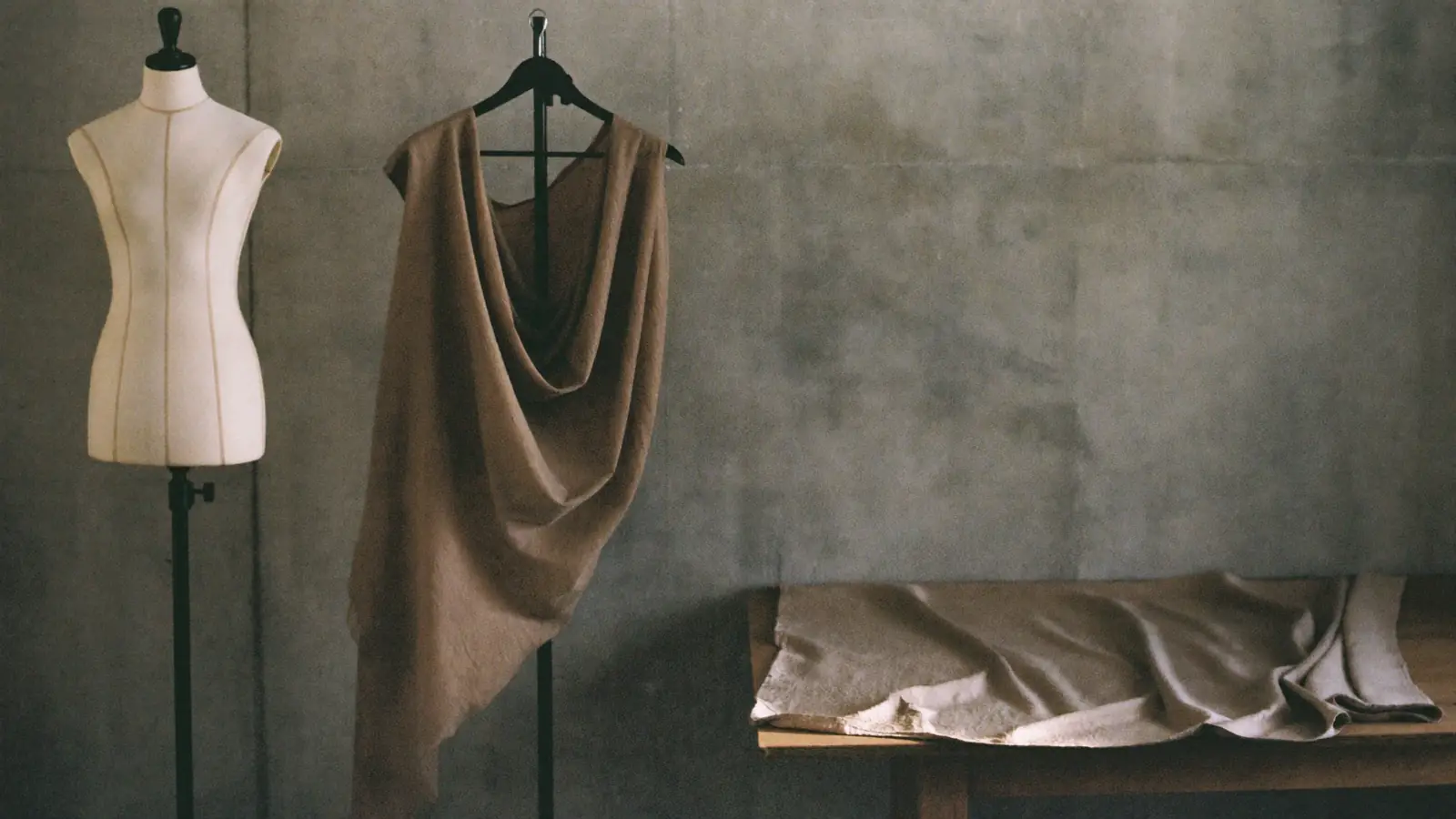Three lengths of cloth hanging side by side against raw concrete, one draped on a stand, one flat on a table, one still folded, hard side light, muted charcoal and bone, the draped one lifting slightly.