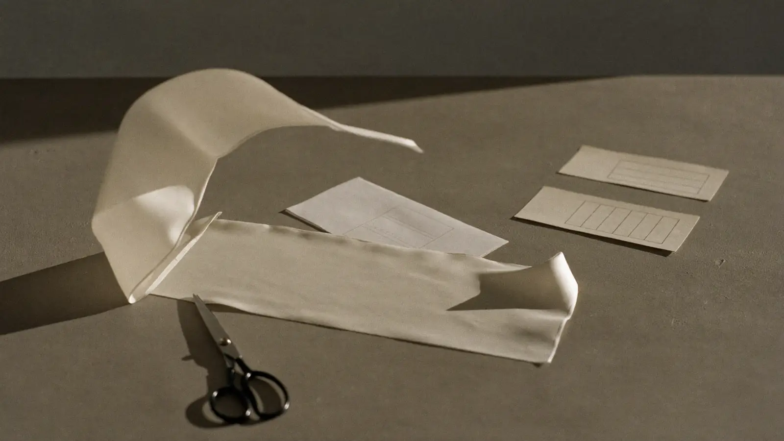 A cut length of ivory silk lying on a bare concrete table beside paper pattern pieces and shears, hard side light, one edge of the silk lifted by a draught, muted bone and grey.