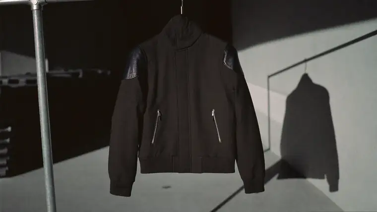 A dark structured stage jacket on a rail in a bare concrete corridor, hard raking side light throwing a long shadow, road cases stacked behind, muted charcoal with one cold highlight on the shoulder seam.