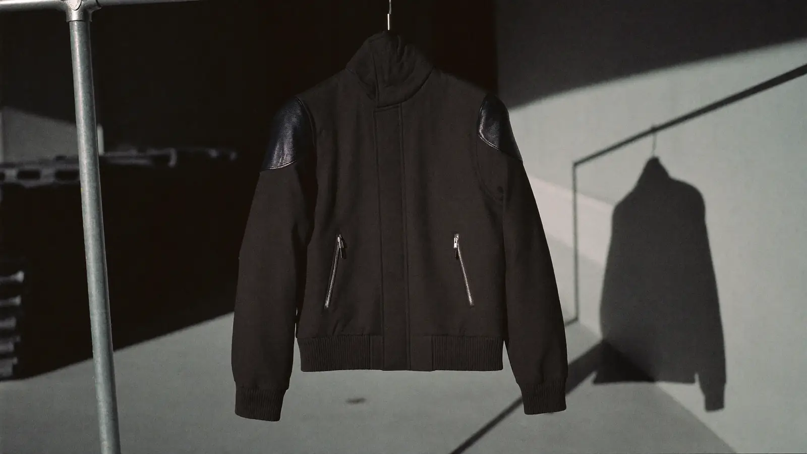 A dark structured stage jacket on a rail in a bare concrete corridor, hard raking side light throwing a long shadow, road cases stacked behind, muted charcoal with one cold highlight on the shoulder seam.