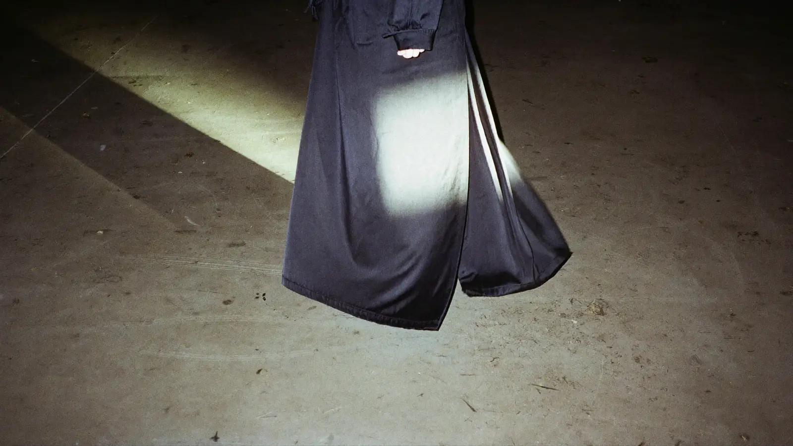 A dark gown lit by one hard flash against bare concrete, a second cold light raking from the side, the hem still moving, muted charcoal and bone with a single bright specular line on the fabric.