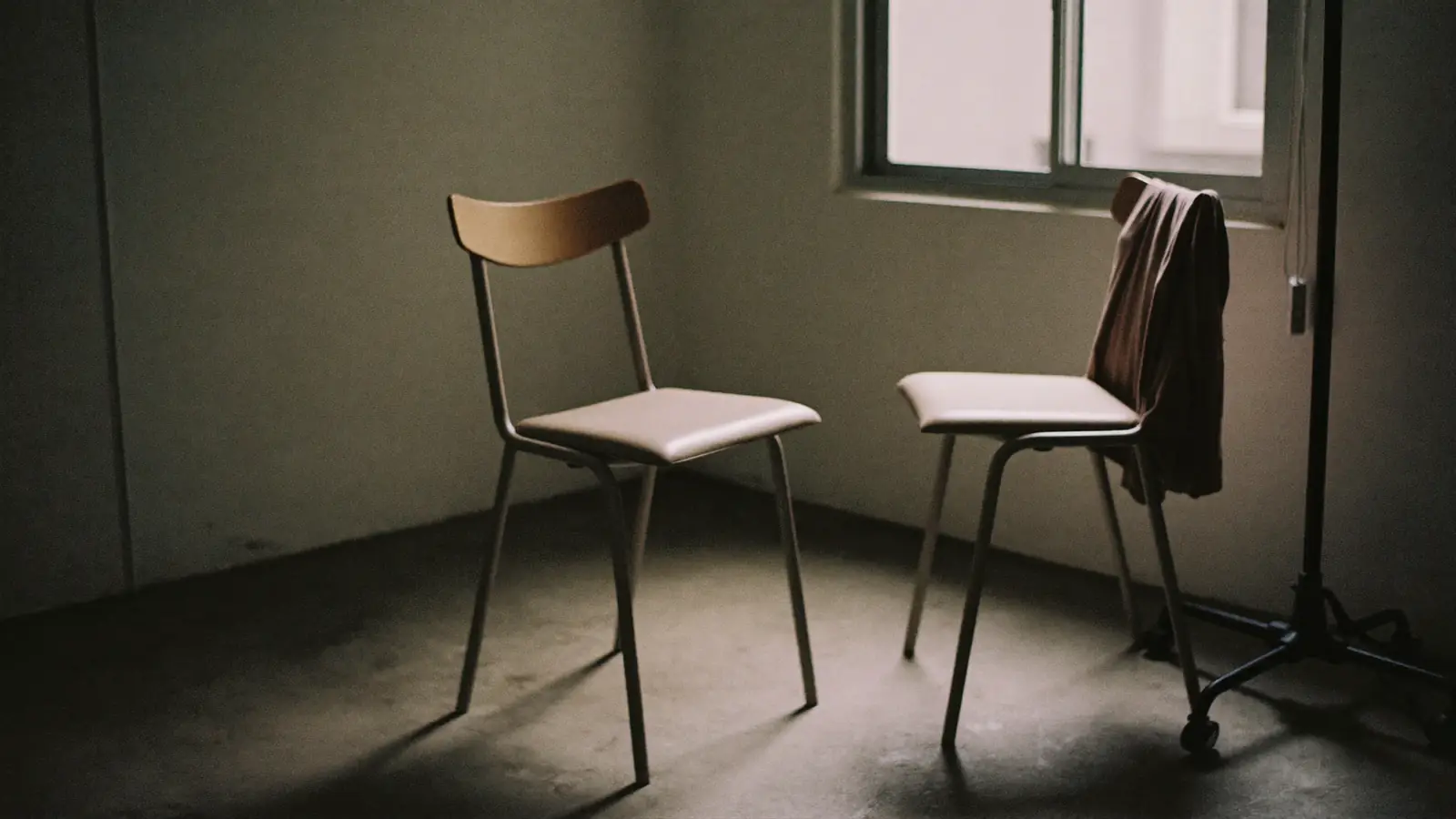Two empty chairs facing each other on a bare concrete floor beside a dress stand, hard side light through a tall window, a length of cloth draped over one chair back, muted bone and grey.