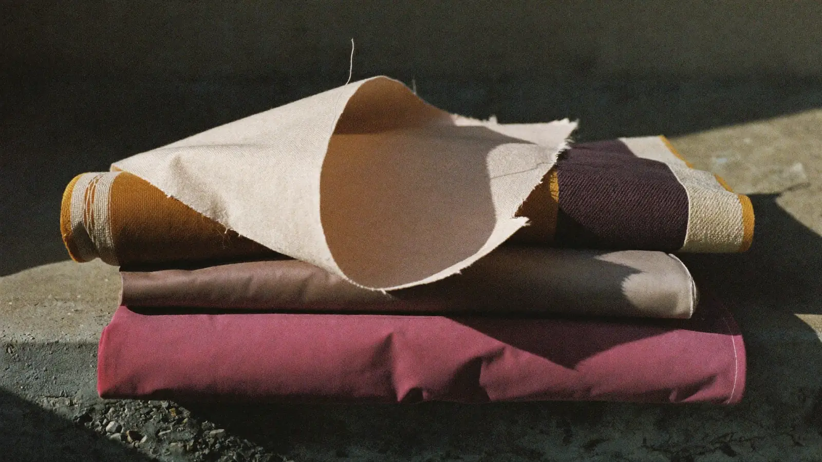 Folded lengths of silk, wool crepe and gazar stacked on a bare concrete ledge, hard side light picking out the weave of the top length, one edge lifting in a draught, muted bone, charcoal and dust pink.