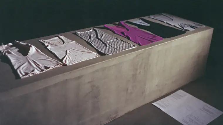 A long bare concrete table with a line-up of garments laid flat in sequence, hard side light from one end, a paper line sheet weighted down at the corner, muted bone, grey and one deep colour.