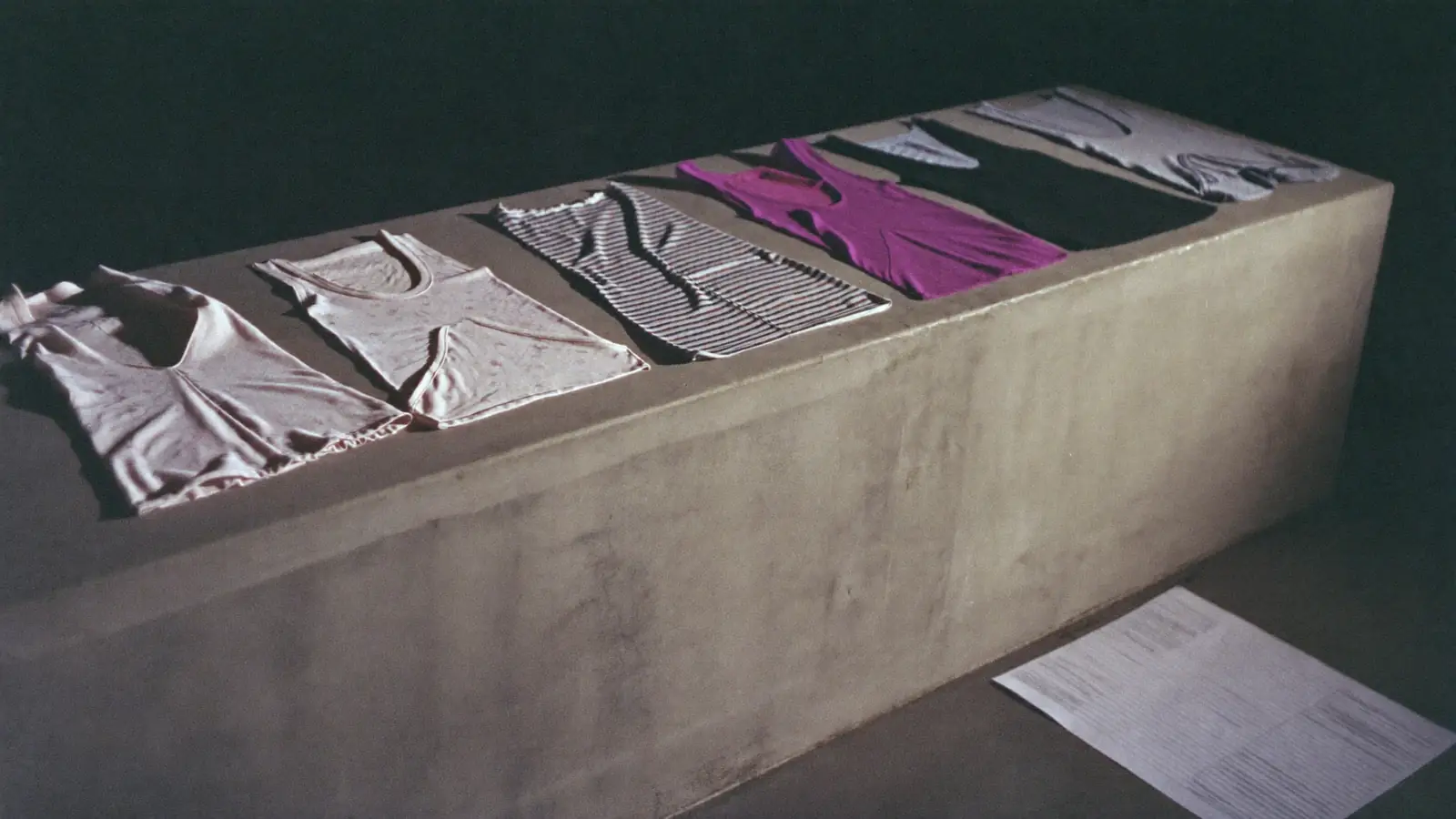 A long bare concrete table with a line-up of garments laid flat in sequence, hard side light from one end, a paper line sheet weighted down at the corner, muted bone, grey and one deep colour.