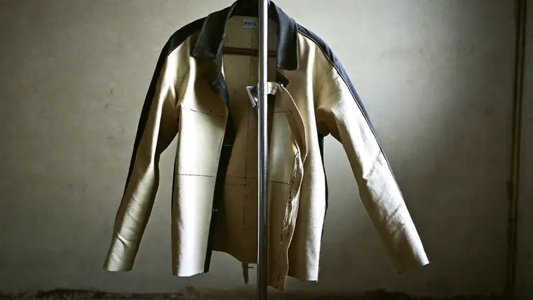 A half-made jacket on a tailor's stand in a bare concrete room, one canvassed front open to show the internal structure, hard side light across the shoulder, muted charcoal and bone, chalk lines visible.