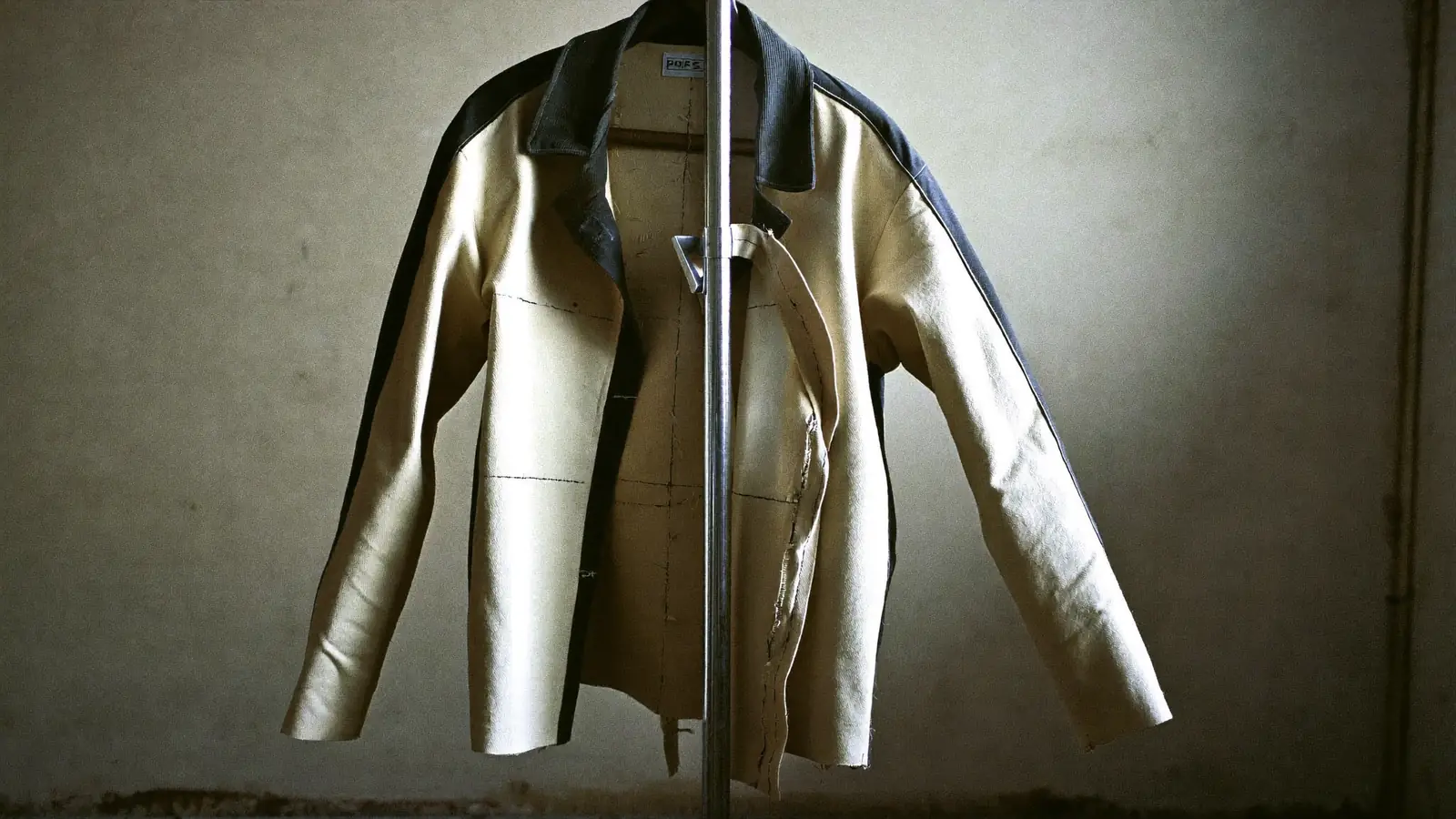 A half-made jacket on a tailor's stand in a bare concrete room, one canvassed front open to show the internal structure, hard side light across the shoulder, muted charcoal and bone, chalk lines visible.