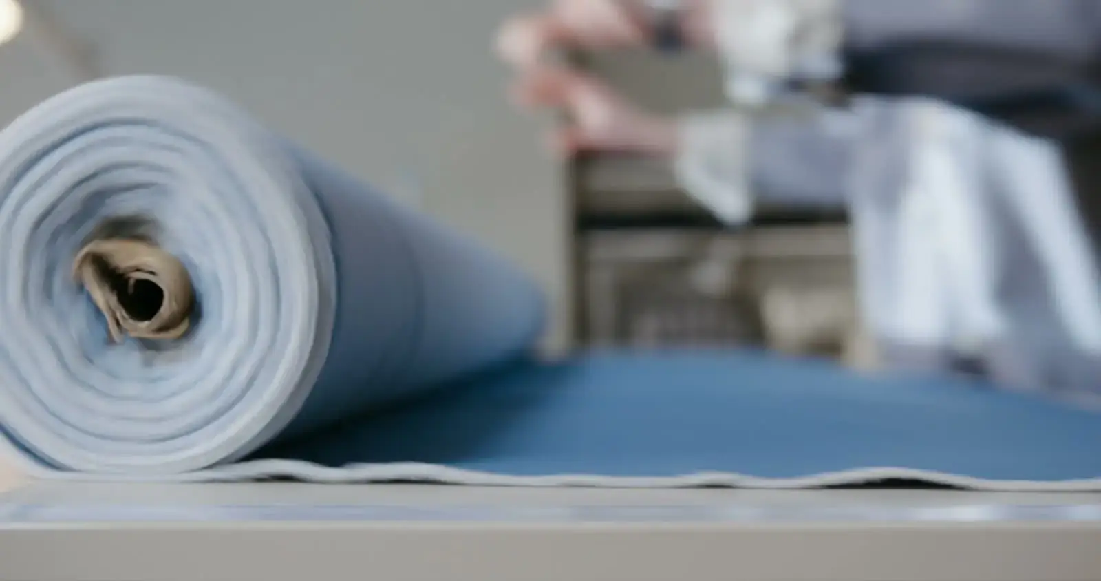 A bolt of pale cloth being unrolled across a cutting table